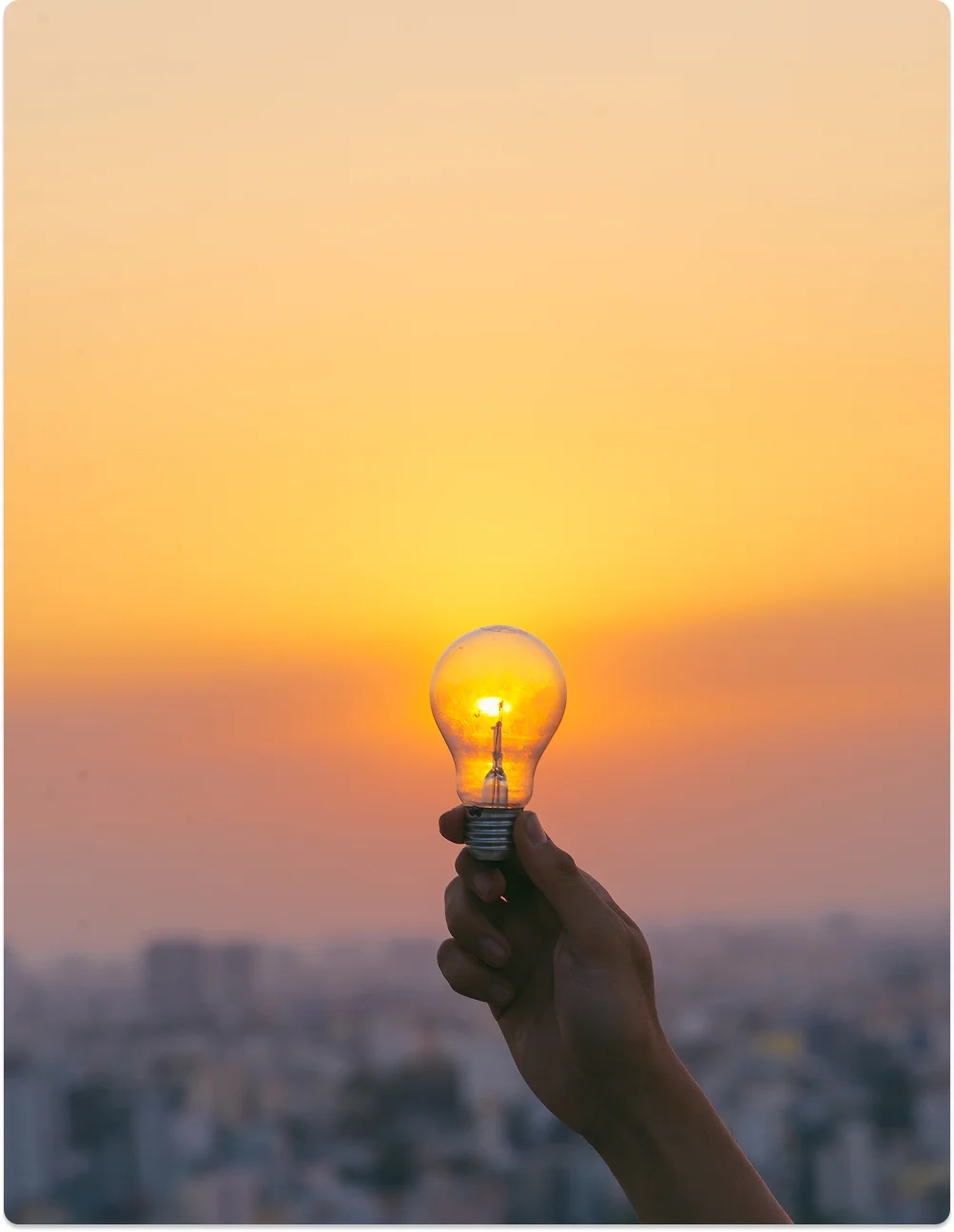 Mano sosteniendo una bombilla encendida frente a un atardecer, simbolizando creatividad, inspiración y claridad en Be MEE Mentoría.